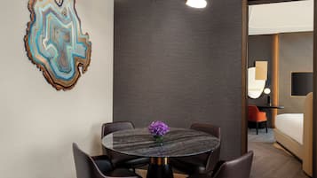 A modern hotel room with a round dining table, leather chairs, a decorative wall piece, and a view into the adjacent room.