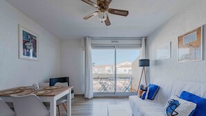 A modern apartment with a dining area, a sofa with decorative pillows, a ceiling fan, and a view through a sliding glass door. - Marine de port, boats and relaxation (Canet-en-Roussillon)