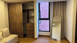 Ein Zimmer mit Holzboden, einem weißen Sofa, einem Holzschrank, einem mit Vorhängen versehenen Fenster und einem Spiegel.