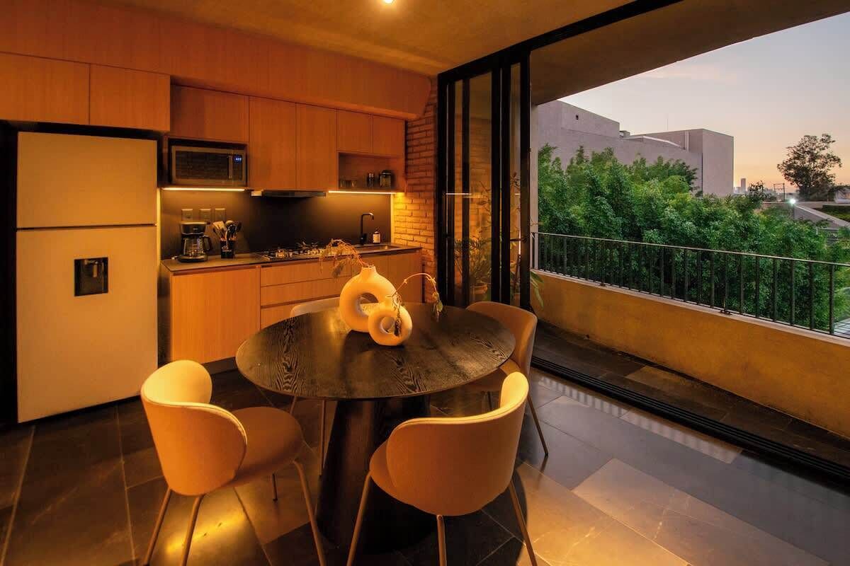 A modern kitchen with a dining area, large windows, and a view of greenery and buildings.