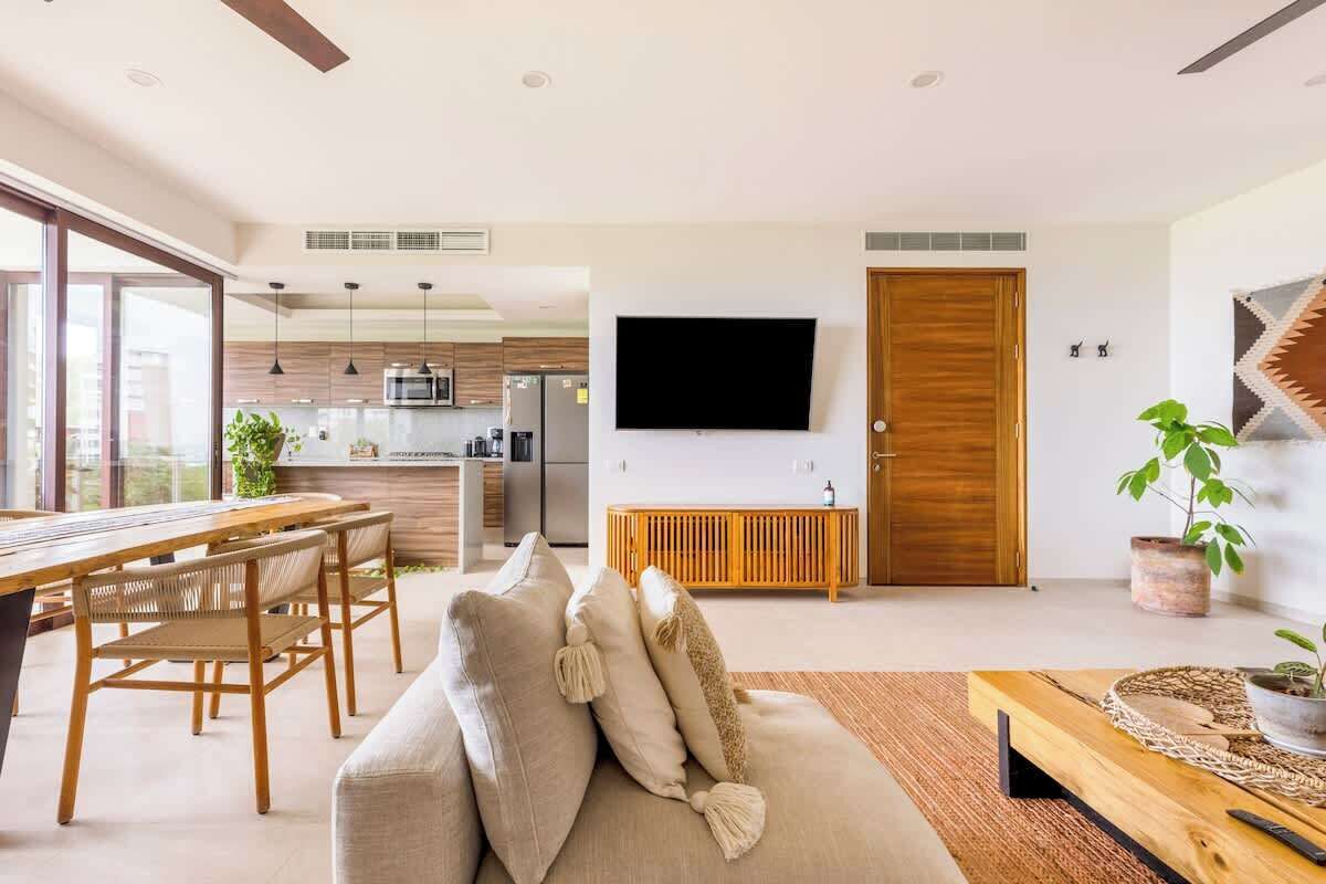 A modern living room with a sofa, dining table, and chairs, a TV mounted on the wall, and a kitchen in the background.