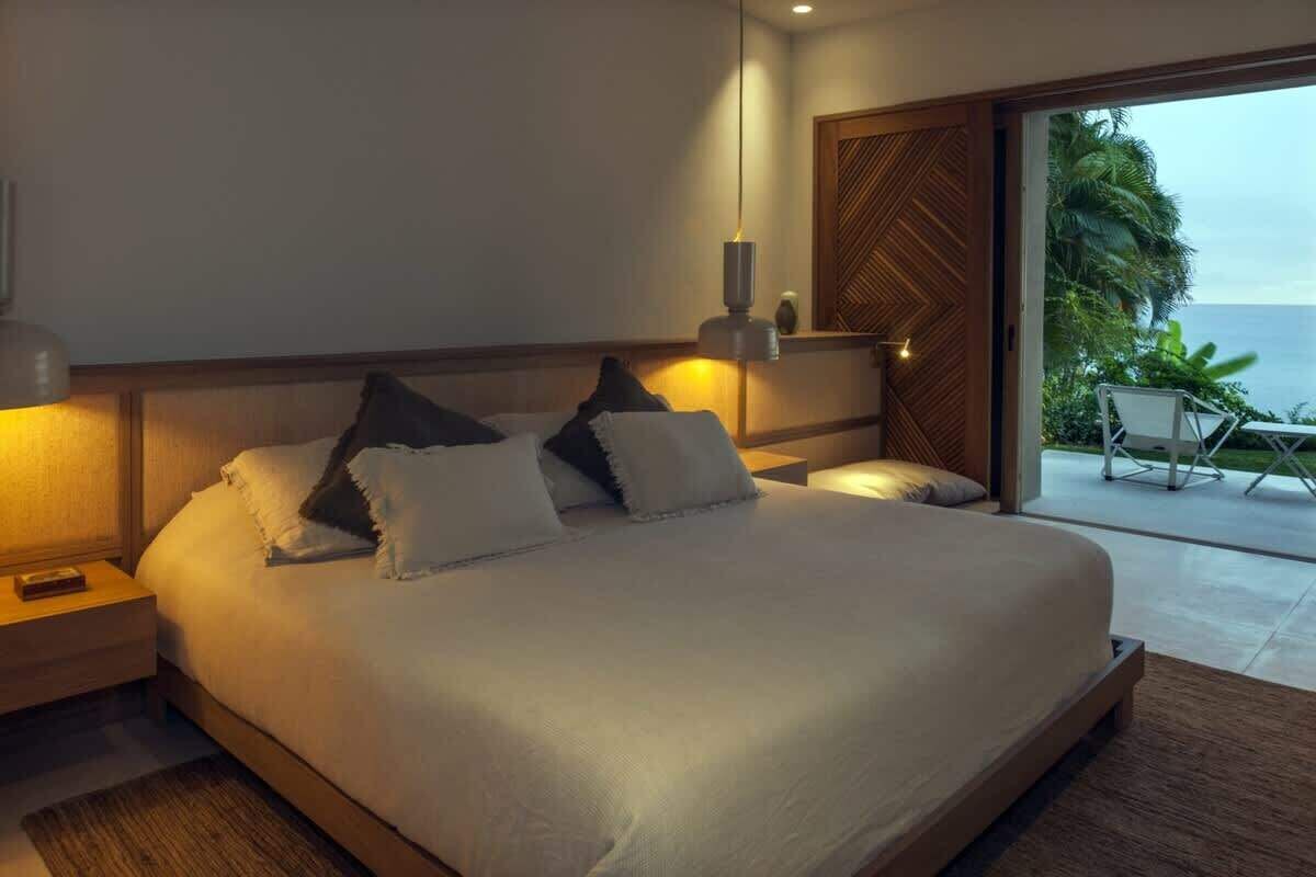 A modern bedroom with a large bed, a nightstand, a wall-mounted lamp, and a view of the ocean through a sliding door.