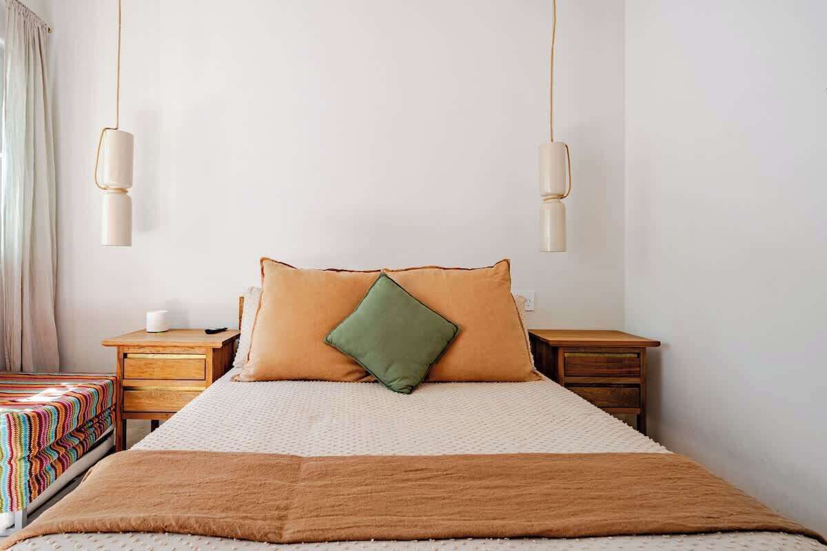 A neatly made bed with a brown blanket, a green pillow, and a beige pillow. There are two bedside tables with lamps and a small bedside table with a lamp.