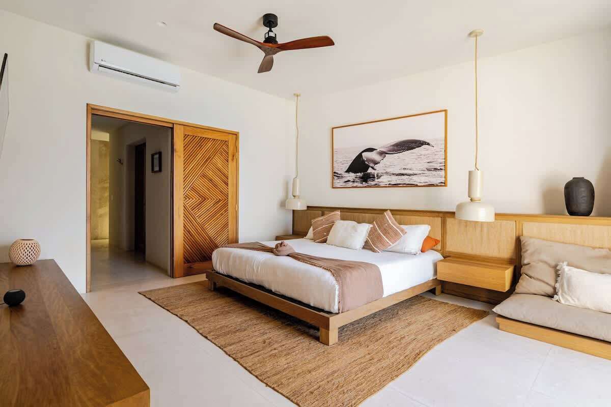 A modern bedroom with a bed, bedside tables, a sofa, a ceiling fan, a framed picture, and a door leading to another room.