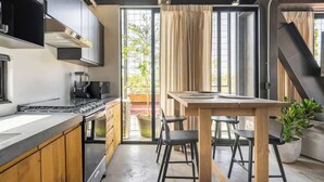 Een moderne keuken met een fornuis, oven en koffiezetapparaat, een eethoek met een tafel en stoelen, en een groot raam met gordijnen.