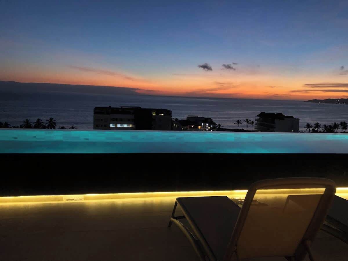 A poolside view with a sunset, a lounge chair, and buildings in the background.