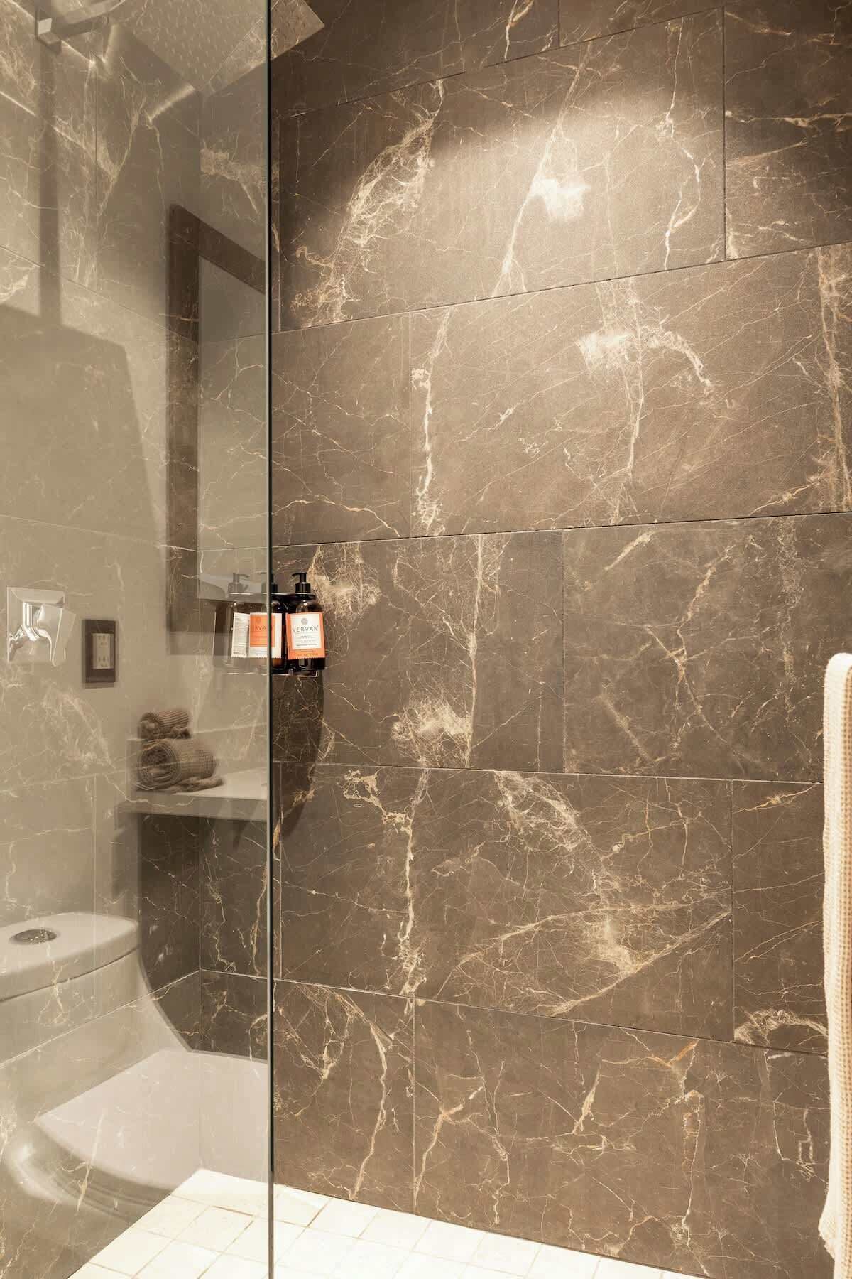 A modern bathroom with a glass shower enclosure, marble walls, and a visible bottle of shampoo.
