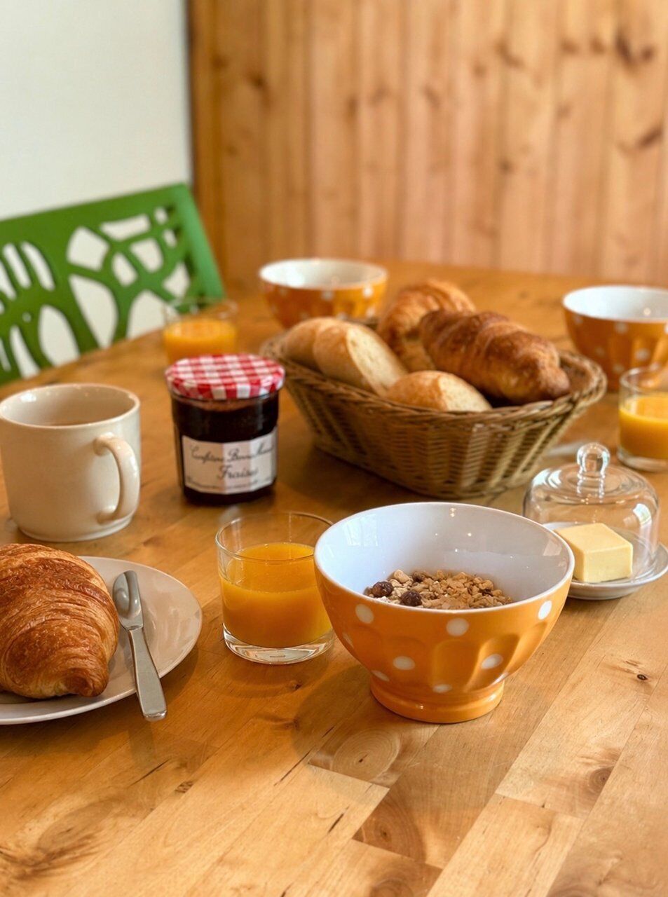 Desayuno con cruasanes, una cesta de pan, un tarro de mermelada, un tazón de cereal, un vaso de jugo de naranja y una taza de café sobre una mesa de madera.