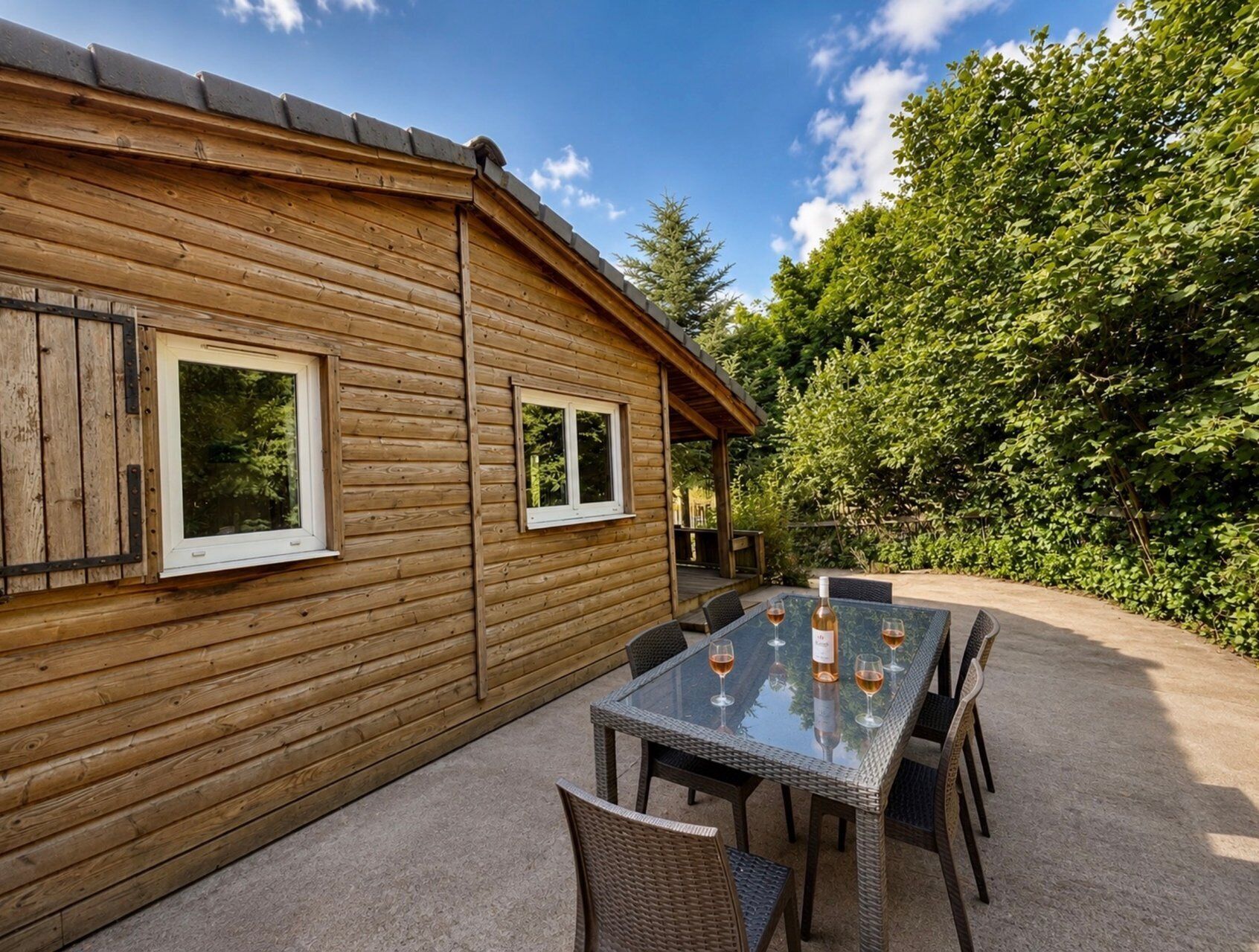 A wooden cabin with a patio area featuring a glass-top table and chairs, surrounded by greenery.