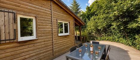 A wooden cabin with a patio area featuring a glass-top table and chairs, surrounded by greenery.
