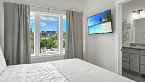 Ein Schlafzimmer mit Bett, einem Fenster mit Vorhängen, einem Fernseher an der Wand und einem Badezimmer mit Waschbecken und Spiegel.