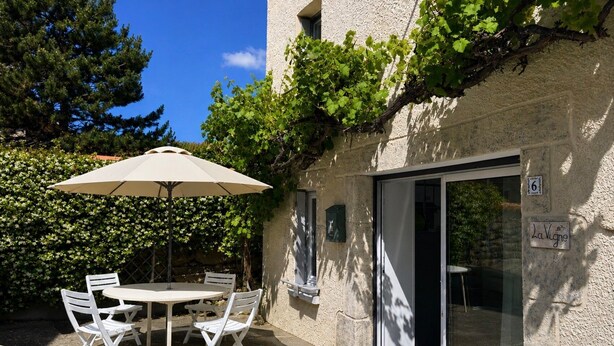 Eine Terrasse im Freien mit Tisch und Stühlen, einem großen Sonnenschirm und einem Gebäudeeingang mit Glastür.