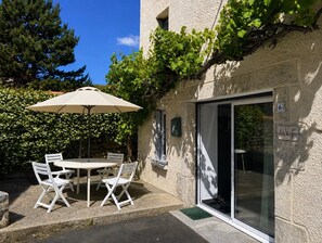 Un patio al aire libre con una mesa y sillas, un gran paraguas y la entrada de un edificio con puerta de cristal.