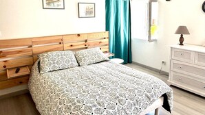 Ein Schlafzimmer mit einem Holzkopfteil, einem Bett mit gemusterten Betttextilien, einem Nachttisch, einer Kommode und zwei gerahmten Bildern an der Wand.
