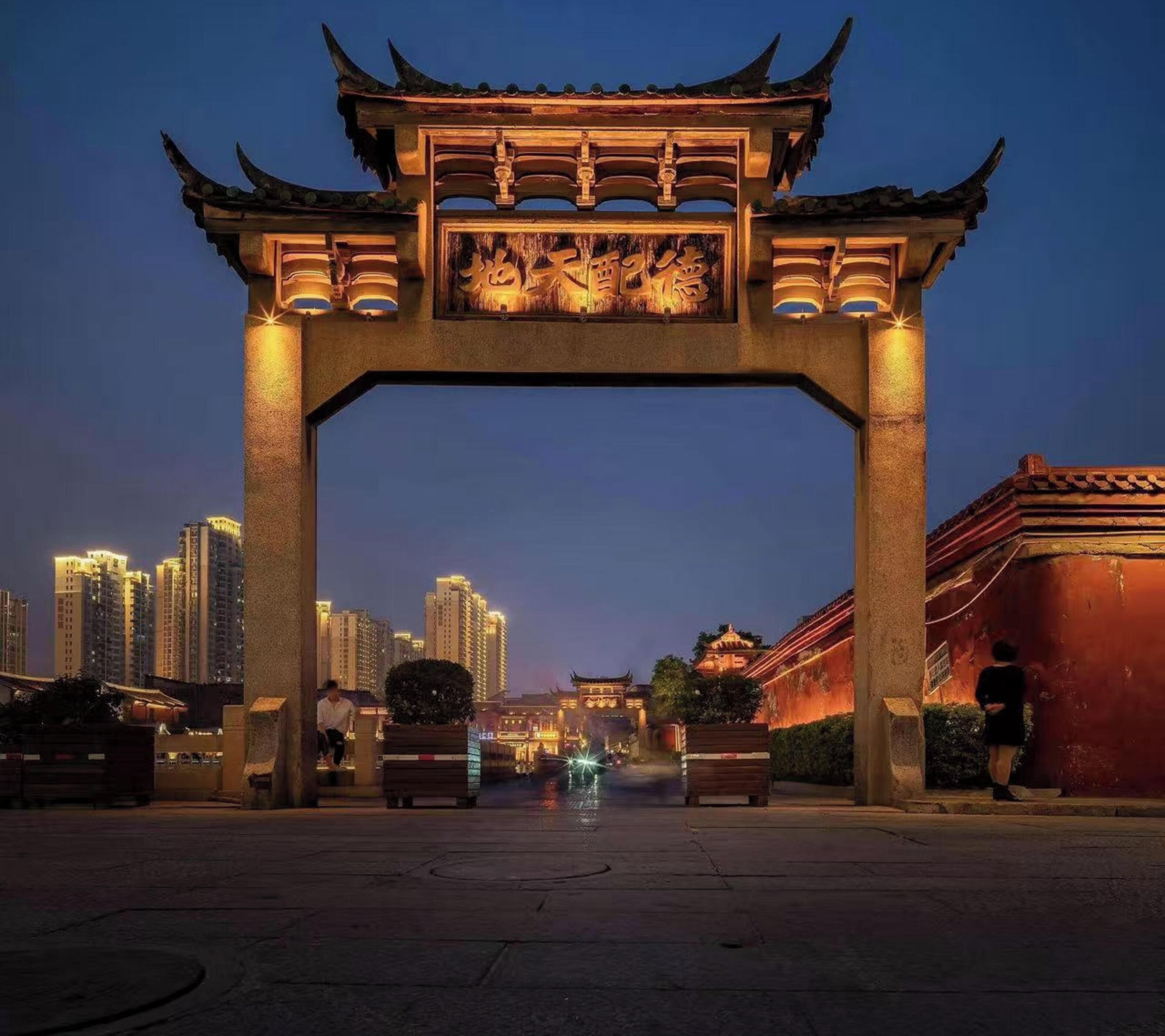 A traditional archway with intricate carvings, leading to a modern cityscape at dusk.