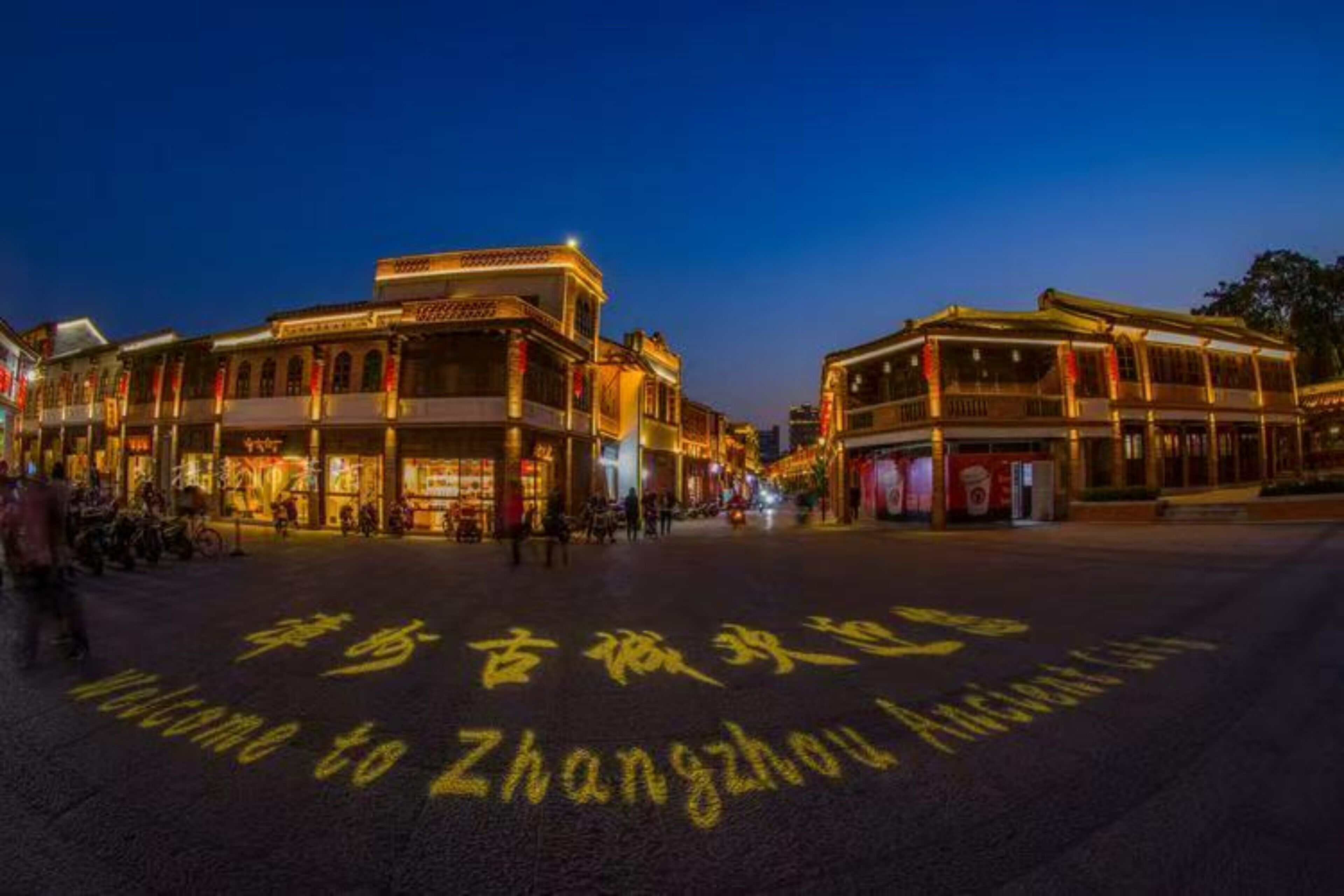 A traditional street scene with illuminated buildings and a welcoming sign in the foreground.