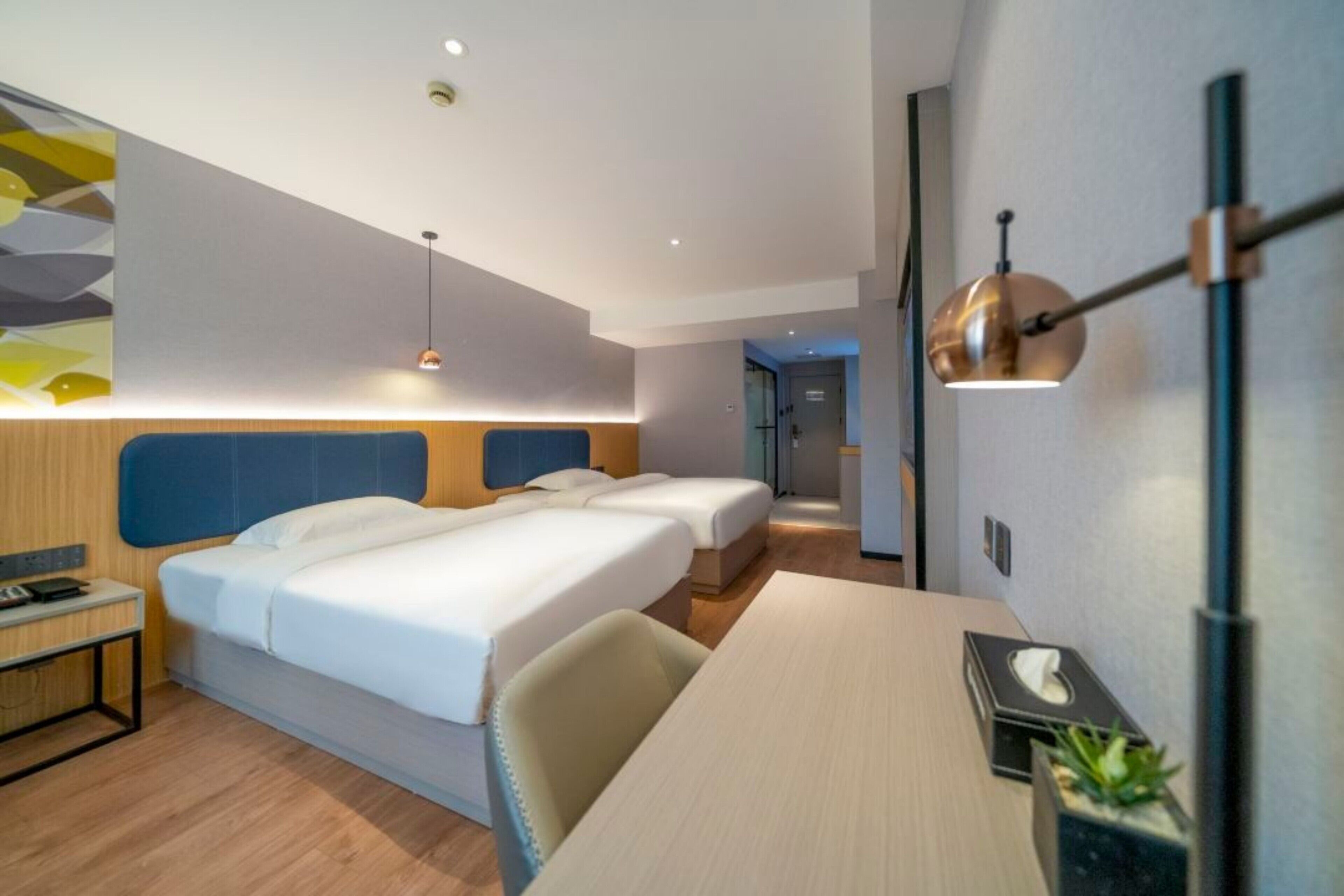 A hotel room with two beds, a desk, a chair, a bedside table, a lamp, and a wall-mounted shelf.