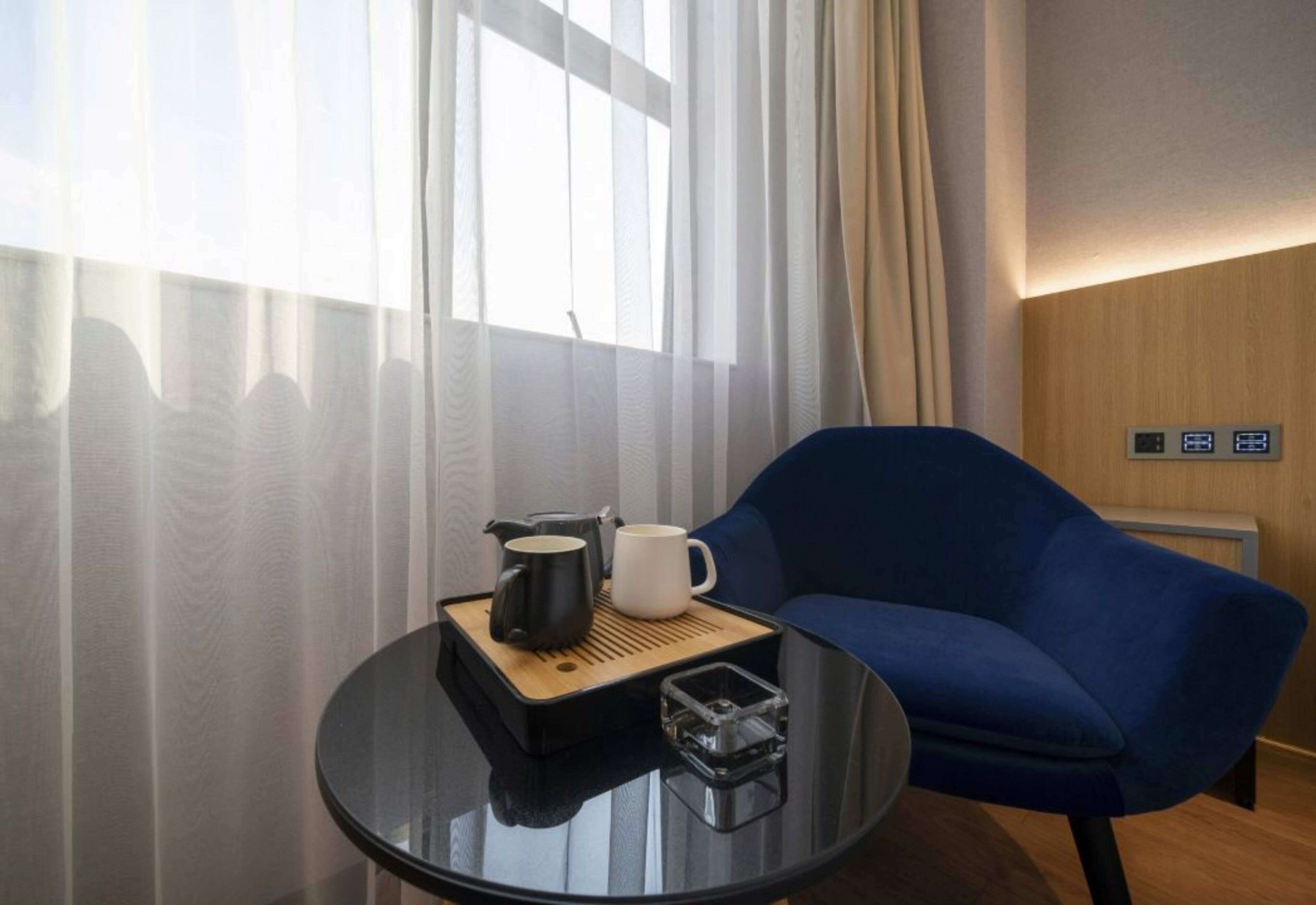 A hotel room with a blue sofa, a small round table with a coffee pot and mug, and a thermostat on the wall.
