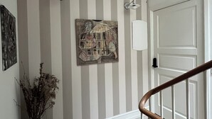 Un couloir avec un papier peint à rayures, un plancher en bois, un vase contenant des fleurs séchées et une porte avec un cadre blanc.