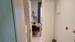 Een hal met een deur links, een bureau met stoel en een plantje op de achtergrond, en een rieten mand op de vloer.