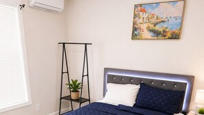 Une chambre à coucher comprenant un lit, une table de chevet, un climatiseur fixé au mur, un tableau et une plante posée sur un support.
