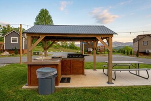 A wooden outdoor barbecue area with a grill, a trash bin, and a picnic table. - Entire Tiny Home Village Near Dollywood Sleeps 120+ (Sevierville)