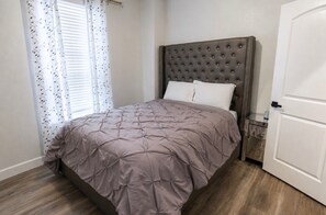 A bedroom with a tufted headboard, a bed with a patterned bedspread, a bedside table, and a door. - Modern 3BR Retreat l Sleeps 7 l 7 Parking Spaces Southeast Houston (Houston)