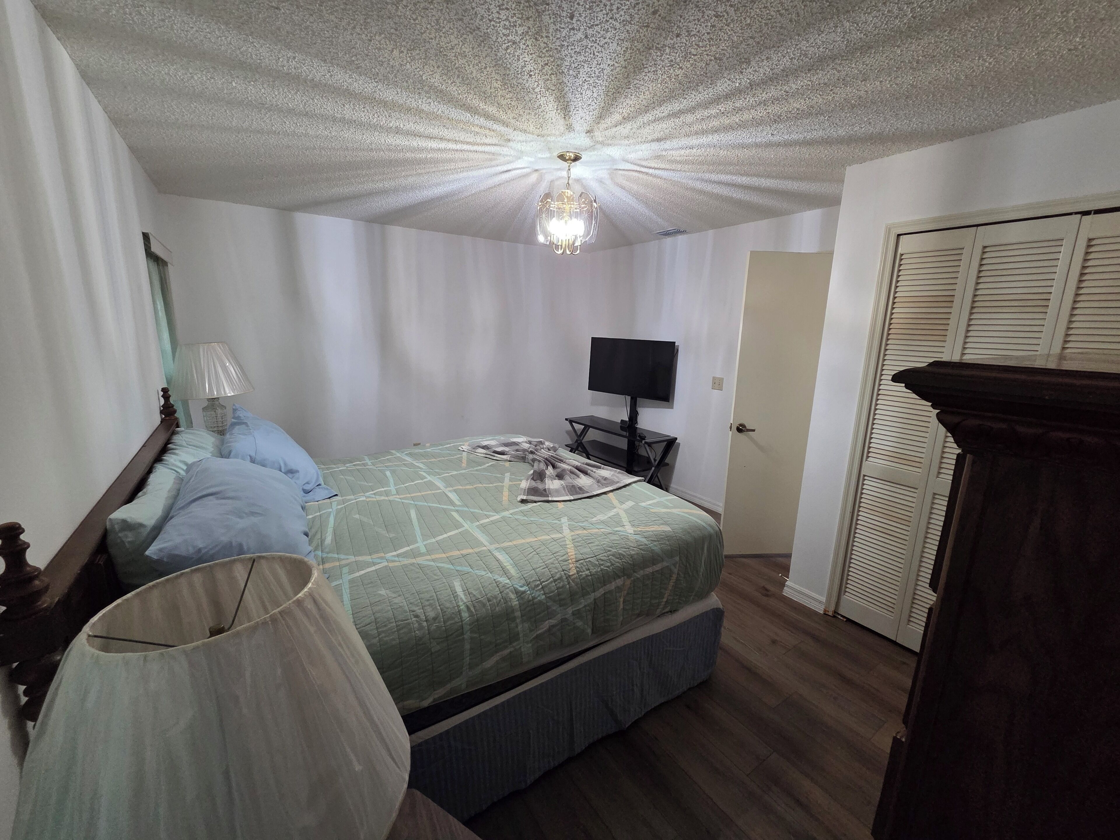 Un dormitorio con una cama, una mesita de noche con lámpara, un televisor y un armario.