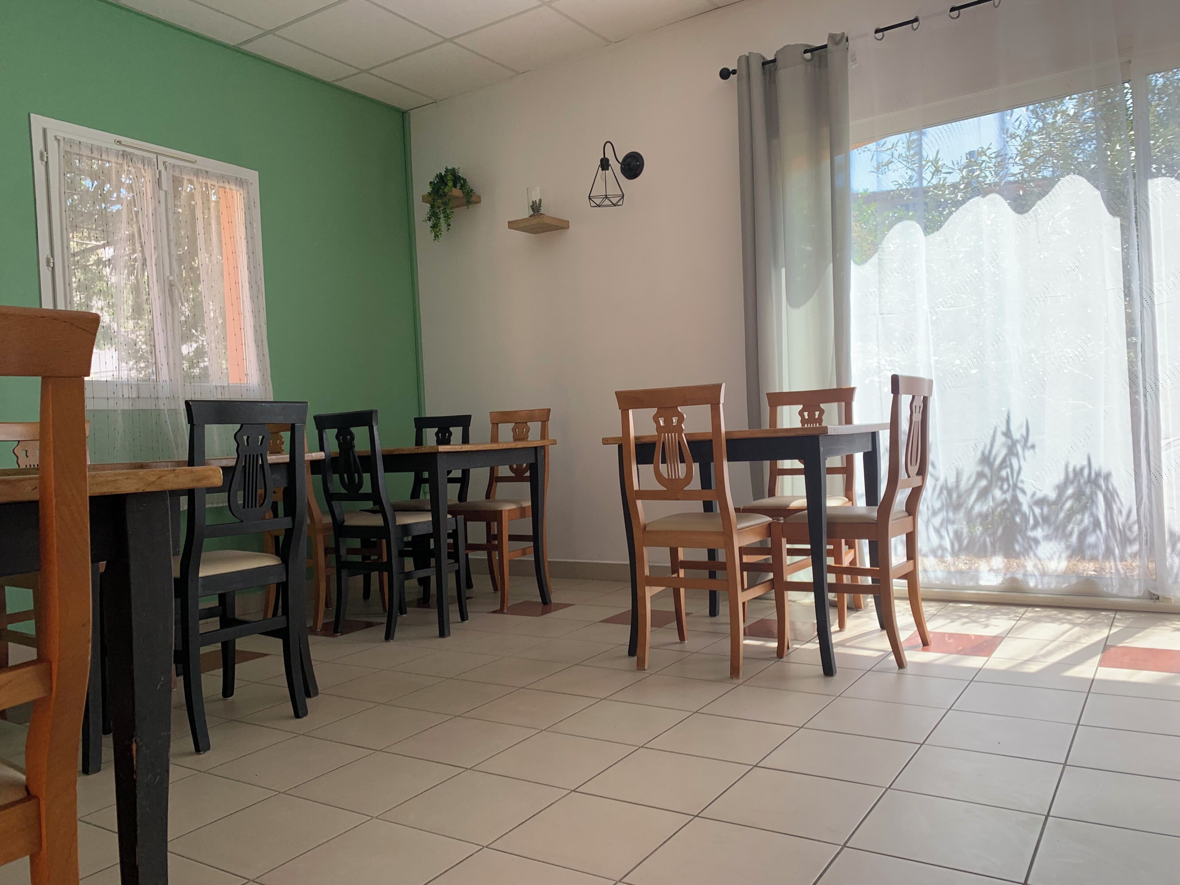 Un espace repas avec des tables et des chaises en bois, un mur végétal, une fenêtre ornée de rideaux légers et une vue sur les arbres à l’extérieur.