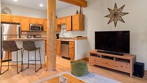 Une cuisine moderne avec des meubles en bois, un four à micro-ondes encastré et un grand téléviseur à écran plat. L’espace détente comprend deux tabourets de bar et un pouf vert.