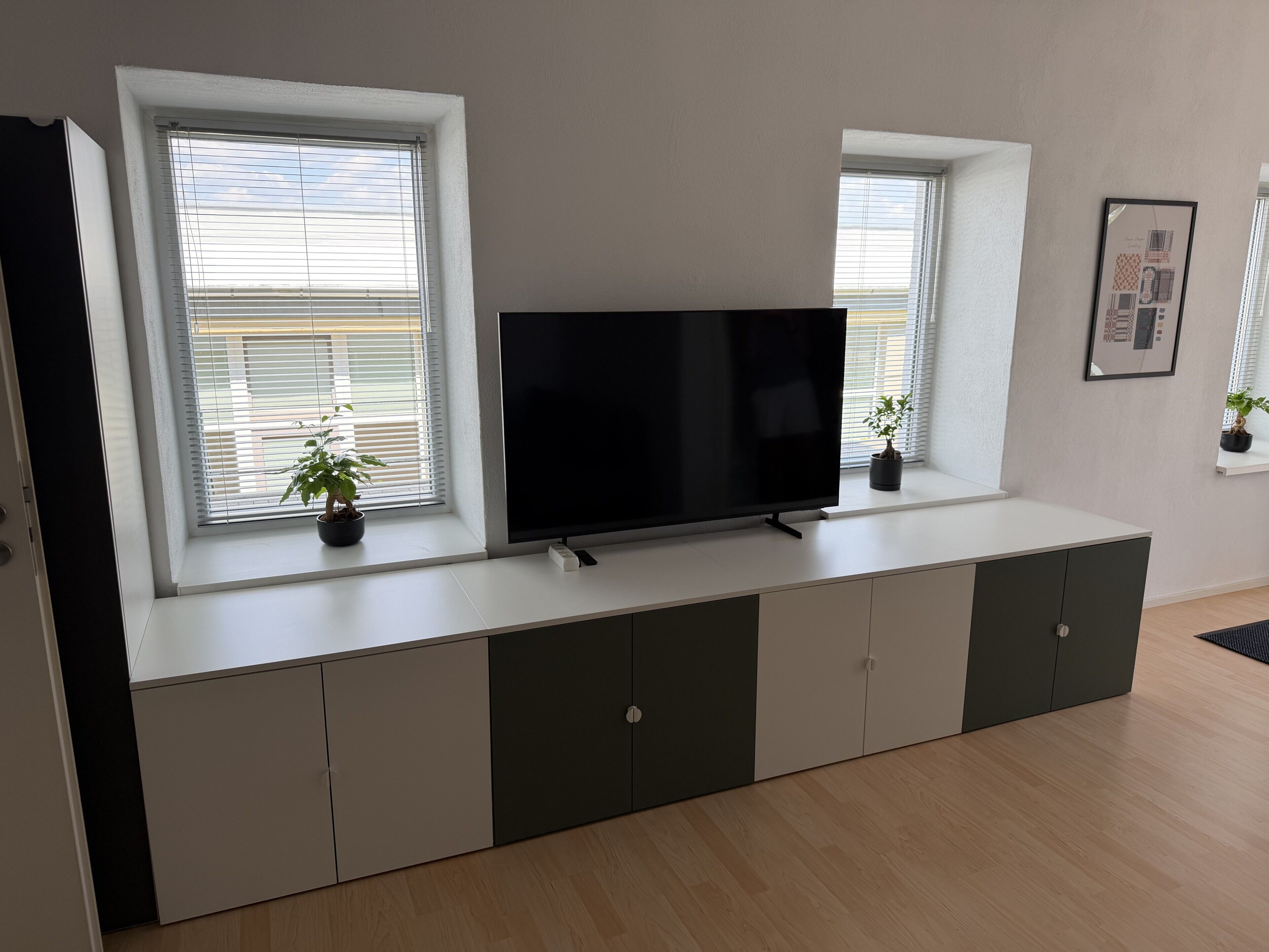 A modern living room with a flat-screen TV on a built-in cabinet, large windows with blinds, and a small potted plant on the windowsill.