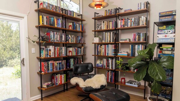 Ein Zimmer mit Holzböchen, gefüllt mit Büchern, einem schwarzen Ledersessel mit flauschigem weißen Kissen, einer Topfpflanze und einem Holzboden.