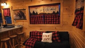 A cozy cabin interior with a wooden wall, a bar area with stools, a couch with a 'STAY COZY' pillow, and a window with a forest scene. - Rustic Tiny Cabin retreat with AC (Union Mills)