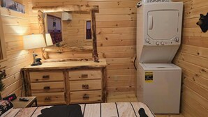 A compact wooden cabin room with a wooden dresser, a mirror, a lamp, a small table with a clock, and a washing machine. - Rustic Tiny Cabin retreat with AC (Union Mills)
