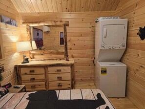 A compact wooden cabin room with a wooden dresser, a mirror, a lamp, a small table with a clock, and a washing machine. - Rustic Tiny Cabin retreat with AC (Union Mills)