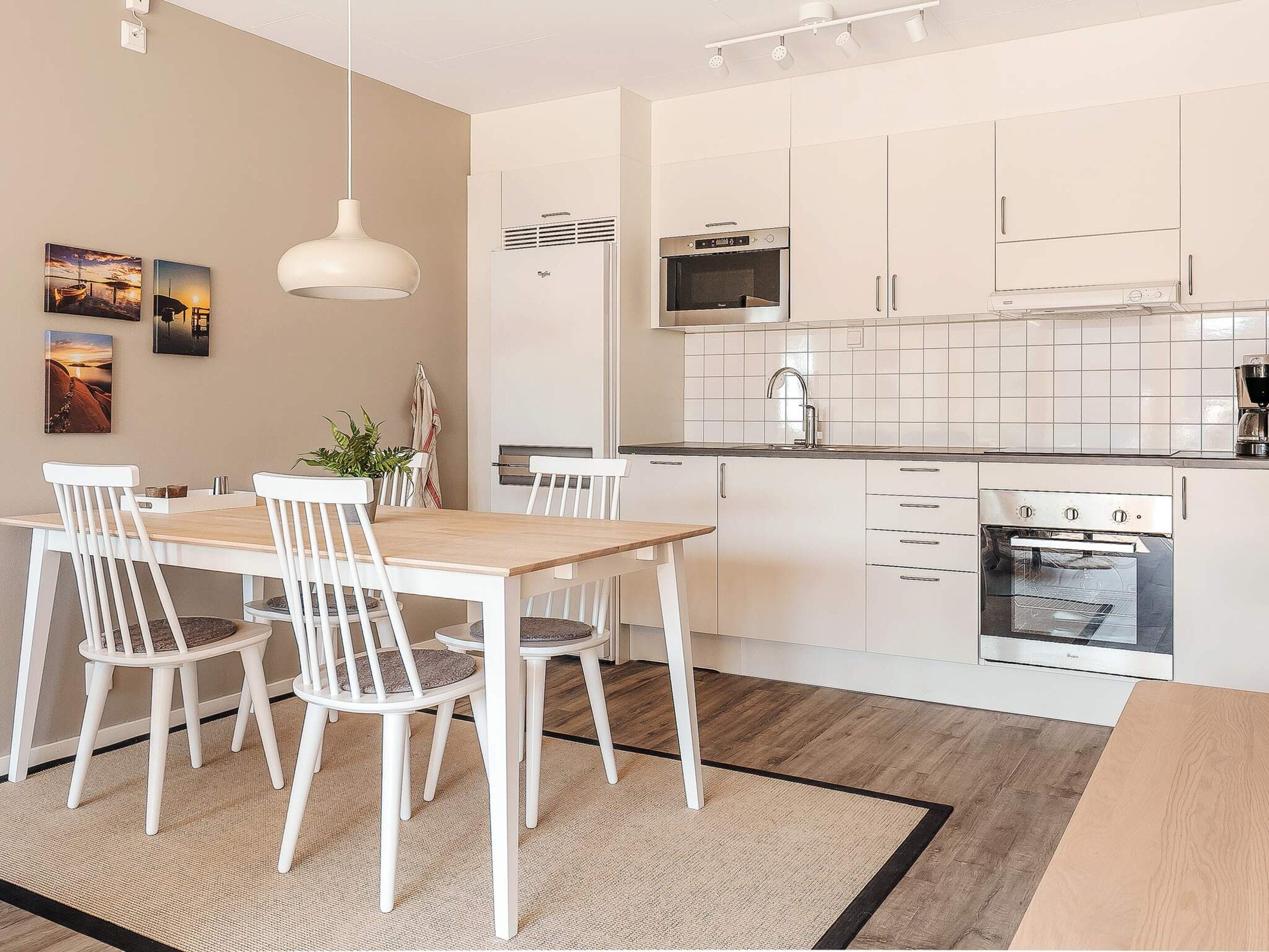 Une cuisine moderne avec des meubles blancs, un four encastré et un coin repas comprenant une table en bois et des chaises blanches.