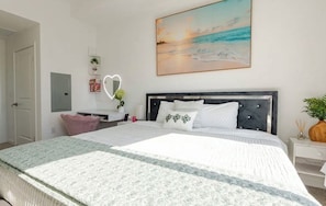 Un dormitorio con cama, mesita de noche, espejo, estante en la pared y un cuadro de una escena de playa.