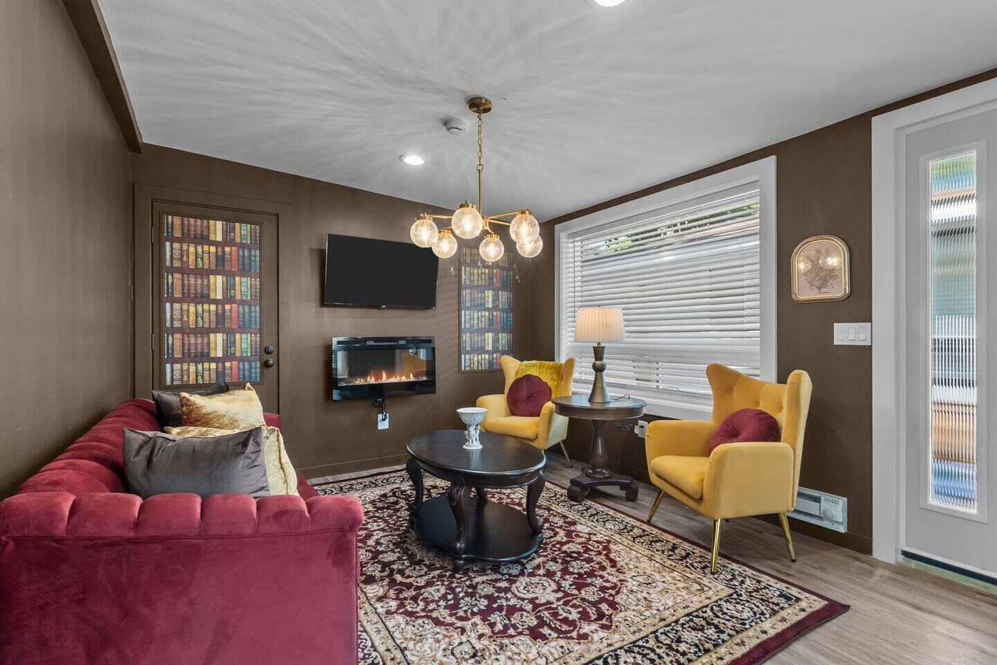 A living room with a red sofa, yellow armchairs, a fireplace, a TV, and a large window with blinds.