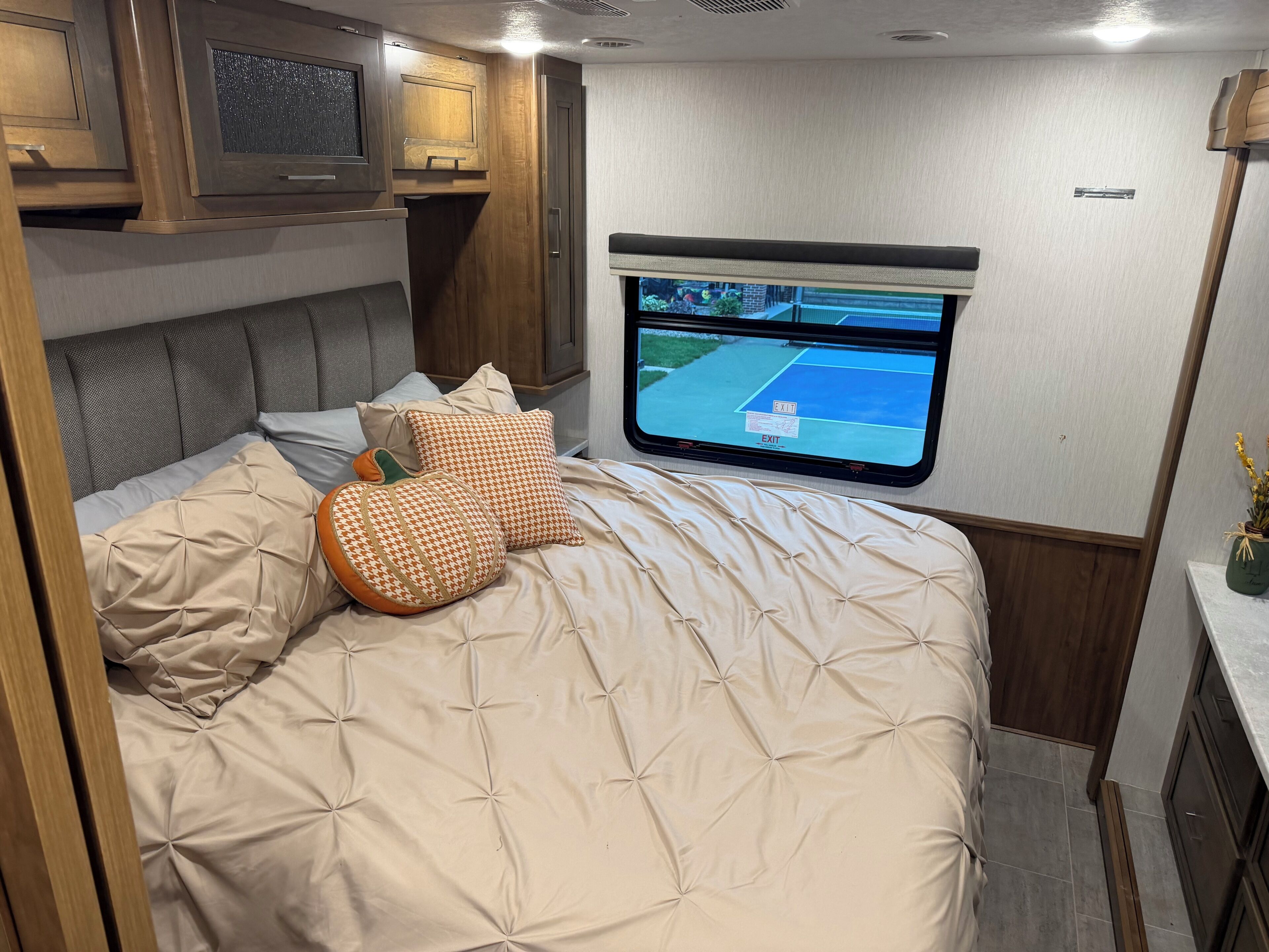 Een bed met een beige sprei, een aan de muur bevestigde televisie en een raam met een 'EXIT'-bordje.