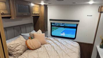 Een bed met een beige sprei, een aan de muur bevestigde televisie en een raam met een 'EXIT'-bordje.