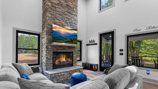 A modern living room with a stone fireplace, grey sofas, and large windows offering outdoor views.