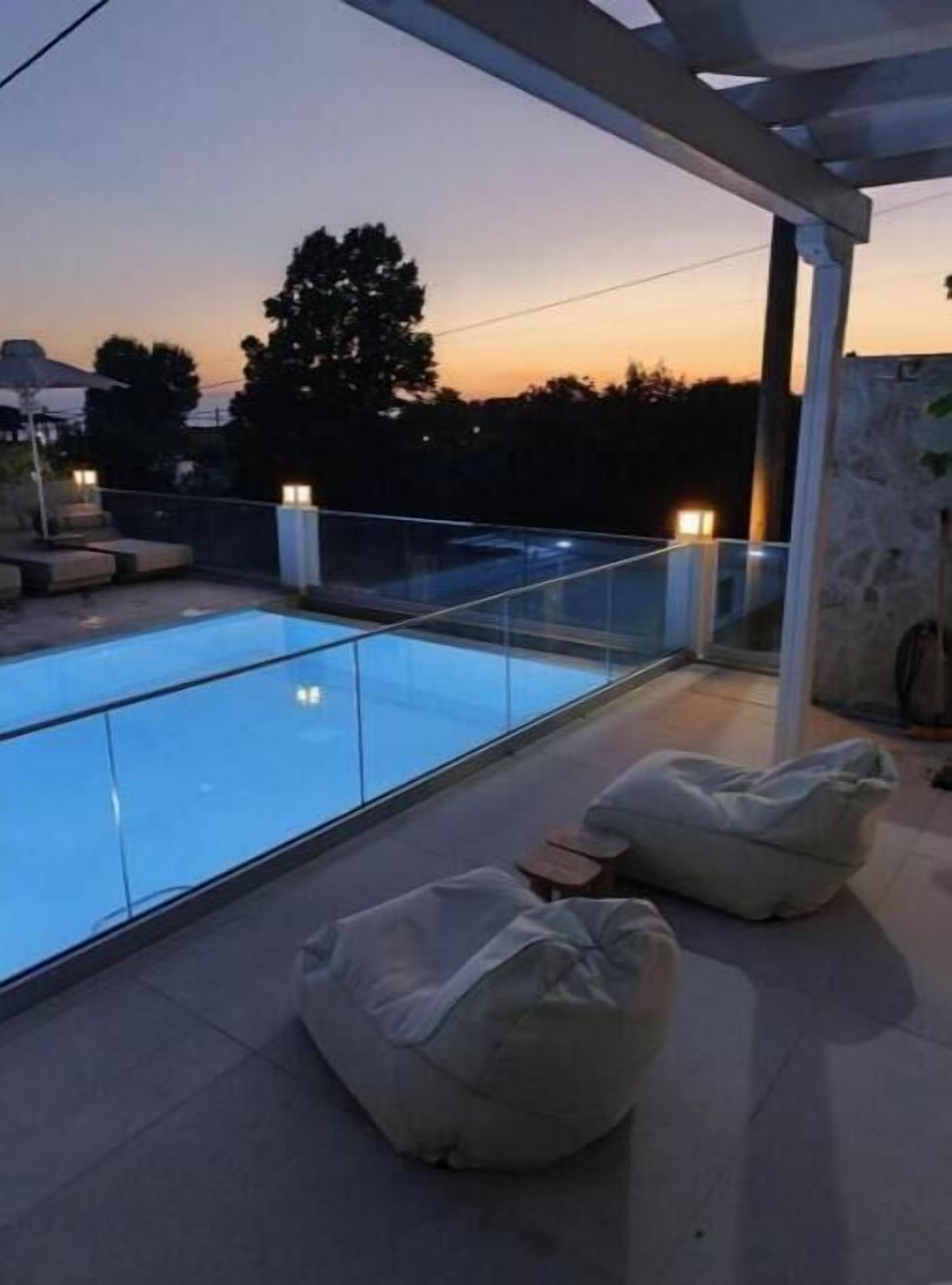 A rooftop pool area with glass railings, two bean bags, and a sunset view.