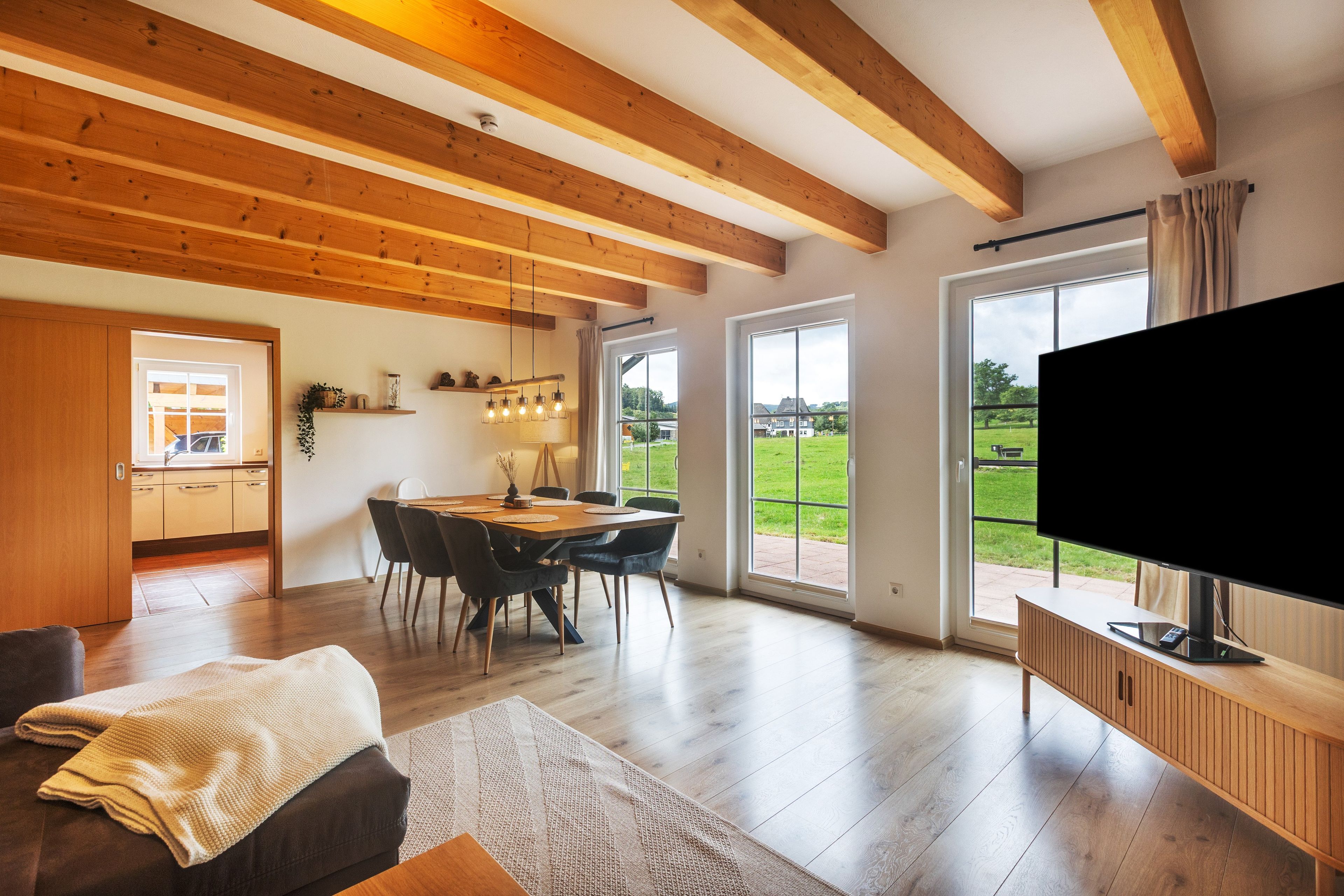 A spacious living area with a wooden ceiling, a dining table with chairs, a flat-screen TV, and large windows offering a view of the outdoors.