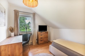 Ein Schlafzimmer mit einem Bett, einem Fernseher auf einer Holzkabine, einem Sessel, einem Schreibtisch, einem Fenster mit Vorhängen und einer Pendelleuchte.