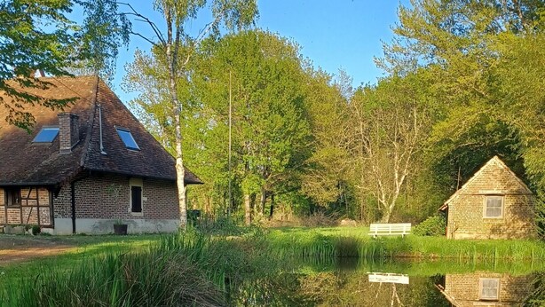 A calm lake with a wooden house and a brick building on the opposite side, surrounded by trees.
