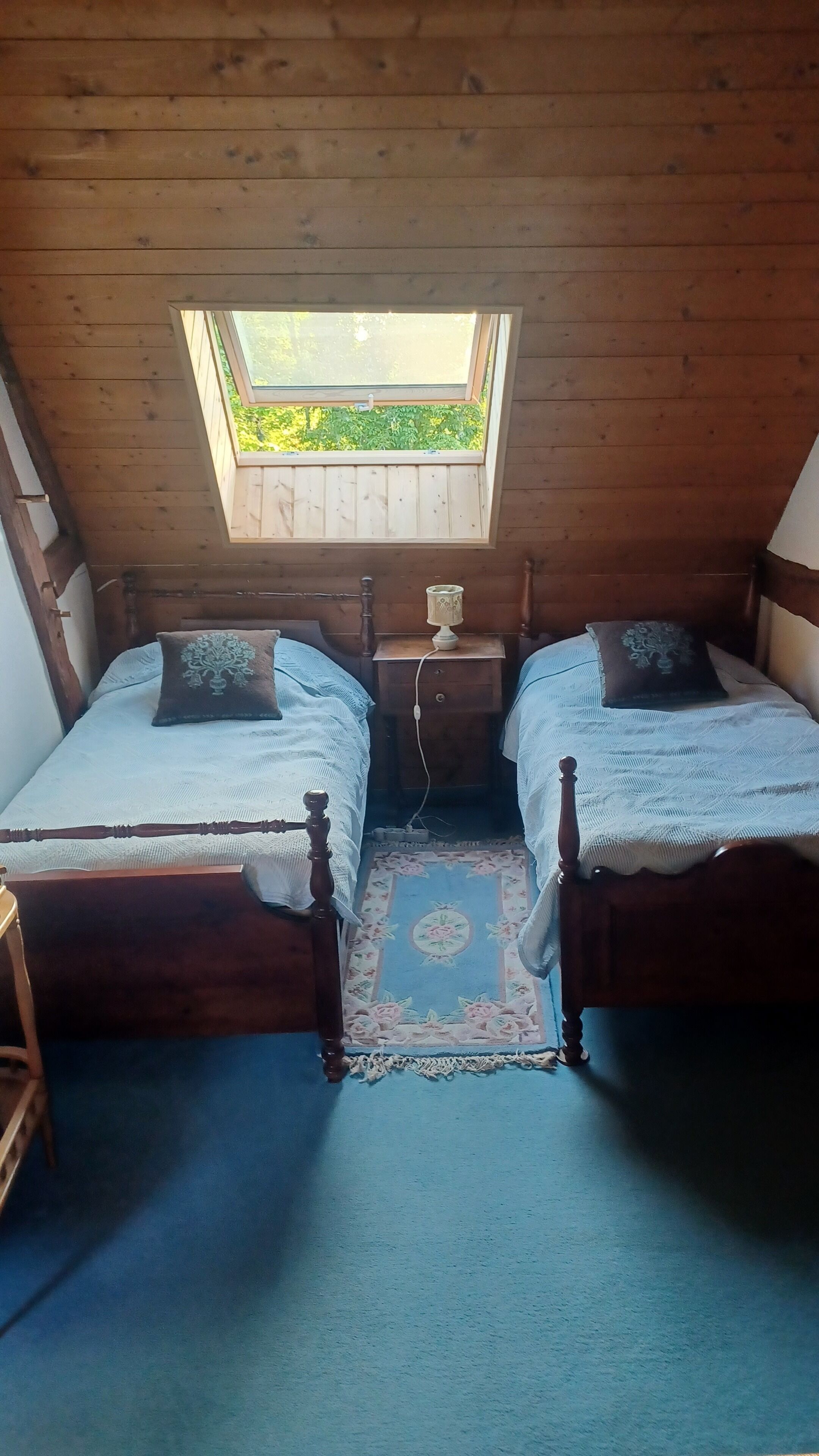 A small, cozy room with two wooden beds, a nightstand, a lamp, and a window with a view of greenery.