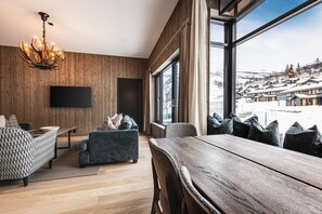 A modern living room with a wooden wall, a chandelier, a flat-screen TV, and a large window with a snowy view. - Ski-in / Ski-out apartment in Hemsedal (Hemsedal)