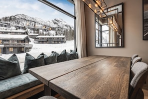 Una mesa de madera para comer con bancos, un ventanal con vistas a edificios nevados y montañas, y una lámpara moderna.