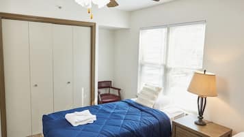 Quarto com duas camas, um ventilador de teto, janela com persianas, uma cadeira e mesa de cabeceira com abajur.