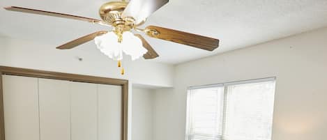 A bedroom with two beds, a ceiling fan, a window with blinds, a chair, and a bedside table with a lamp.